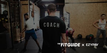 The image captures a dynamic scene inside a modern gym or fitness studio, where a coach is centrally positioned with their back facing the viewer. The word 