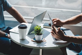 Two people sit at a table with their laptops. Only their hands are visible. 