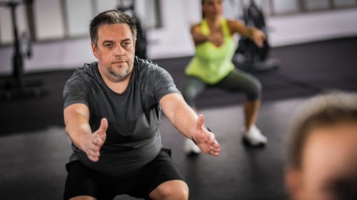 Man in a gym performing a squat with arms extended forward, focused expression. Woman in background doing the same. Bright, active atmosphere.