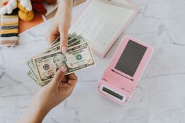 Two hands exchange US dollar bills over a marble table. Nearby are colourful yarns, a notepad, and a pink calculator. 