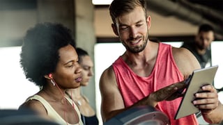 A personal trainer is showing a tablet to a woman at a gym. Both are engaged in conversation, while two other individuals are in the background.