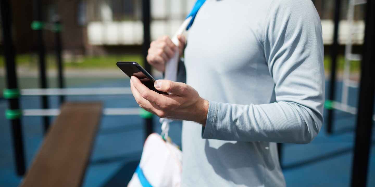 The image captures a moment of focused preparation in an outdoor fitness environment. The individual, dressed in a fitted, long-sleeved, light grey athletic shirt, holds a smartphone in one hand while the other hand adjusts a pair of white wireless earbuds. This suggests a pre-workout routine commonly seen today, where music or guided exercise apps play a key motivational role.
