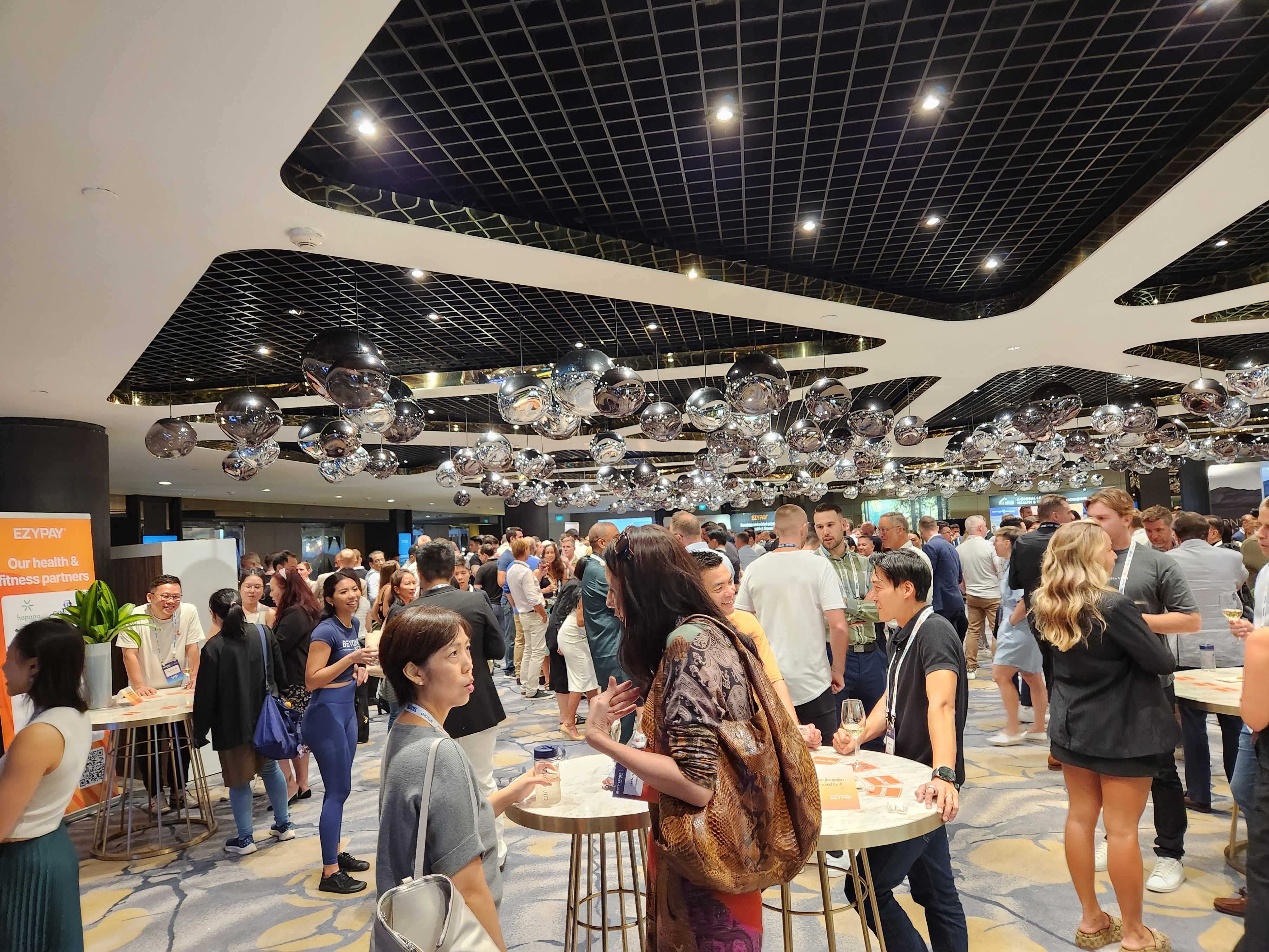 A bustling indoor event space filled with numerous attendees engaged in conversation, surrounded by sleek decor and shiny ceiling installations.