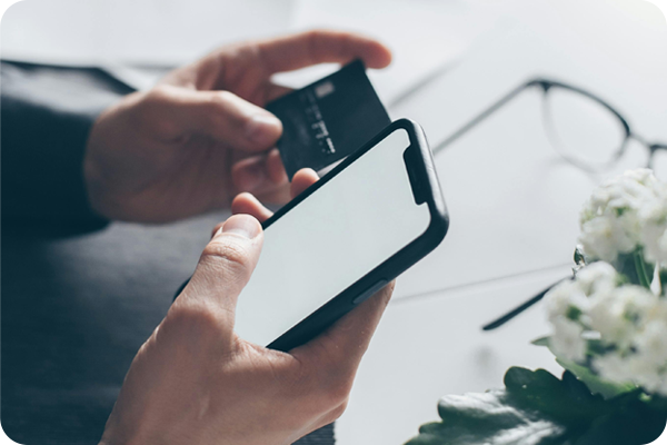 A person holds a smartphone and credit card, suggesting online shopping or mobile payment. Glasses and white flowers are in the blurred background.
