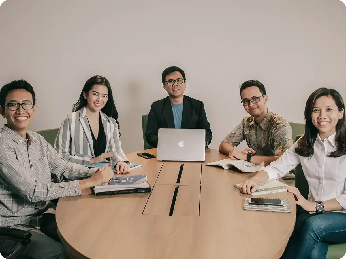 A group of five people sits around a wooden table, smiling. Laptops and books are open, suggesting a collaborative and positive meeting atmosphere.