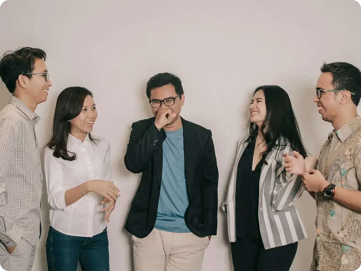 A group of five people stands against a plain background, laughing and smiling. They are casually dressed, conveying a joyful and relaxed atmosphere.