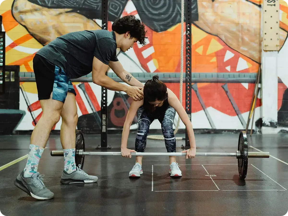 A trainer helps a woman with lifting form in a gym, with a colourful graffiti backdrop. The scene conveys support, focus, and determination.