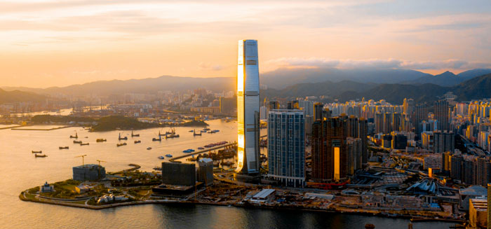 Aerial view of a cityscape at sunset, featuring a tall, illuminated skyscraper amid smaller buildings near a waterfront. Calm, warm tones dominate the scene.
