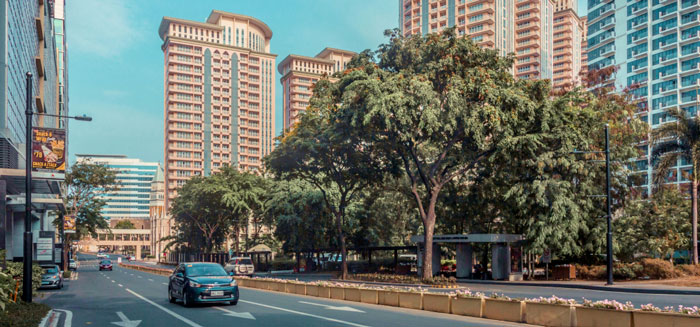 City street with a broad roadway lined by tall, modern apartment buildings and lush green trees. A black car drives down the road under a clear blue sky.