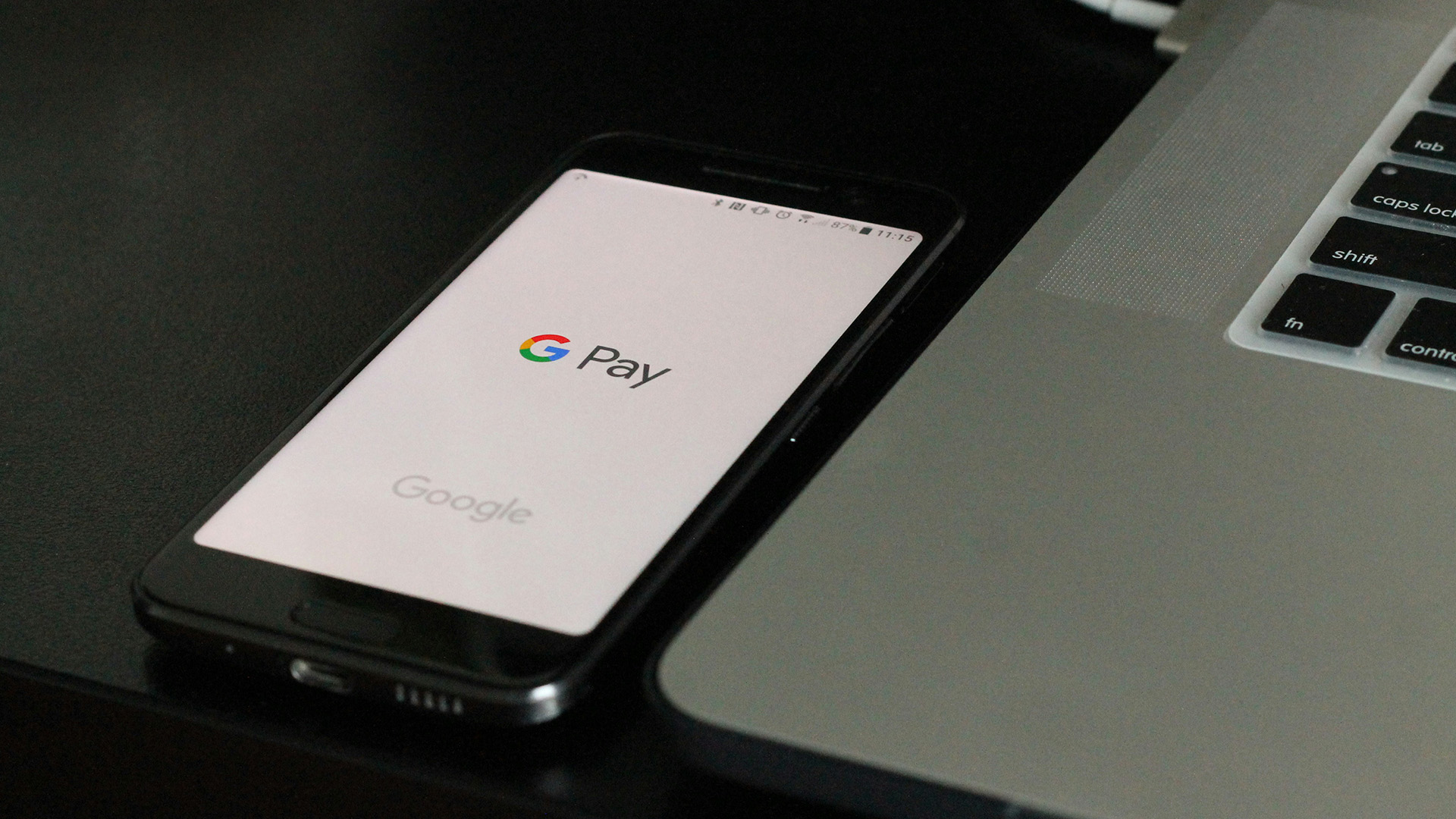 A smartphone displaying the Google Pay logo rests on a dark surface beside a silver laptop. The scene conveys a modern, digital payment setup.