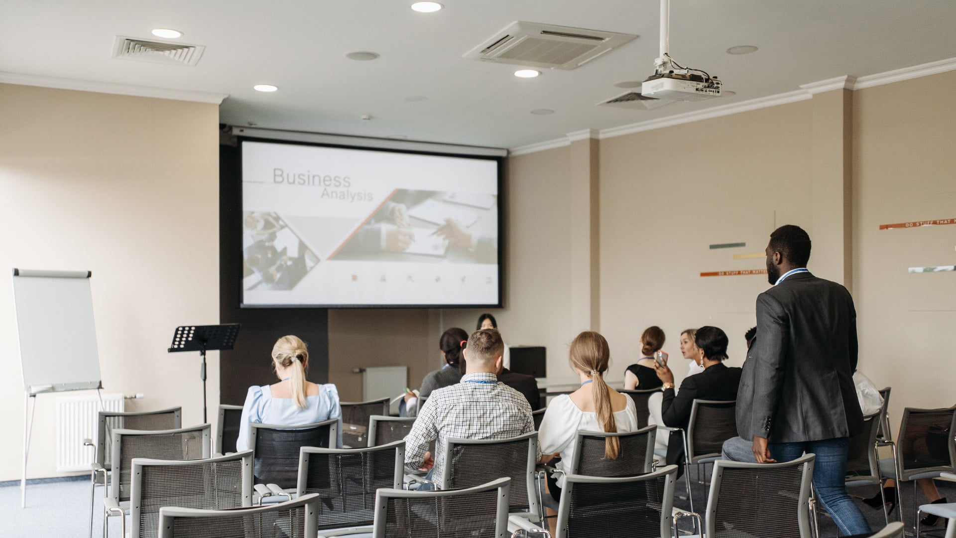 A group of people sits in a conference room, facing a large screen displaying 